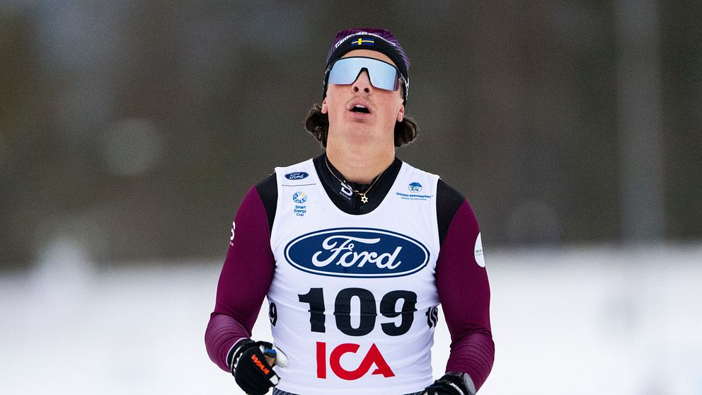 En skidåkare iförd ett tävlingsdräkt med startnummer 109 deltar i ett längdskidåkningslopp. Tävlingsdräkten är märkt med logotyper från Ford och ICA. Skidåkaren är i rörelse över en snötäckt bana med en suddig bakgrund av skog.