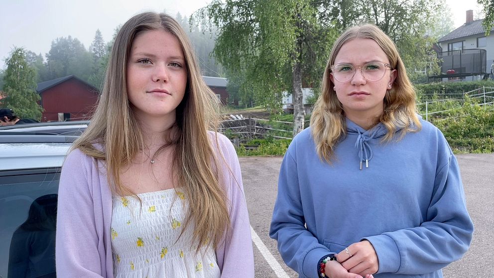 Två tjejer står på en parkering – den ena har långt hår och en lila kofta, varpå den andra bär en blå huvtröja och glasögon.