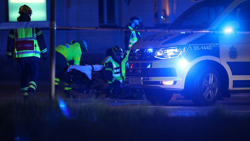 En polisbuss med blåljusen på står bakom en polisavspärrning, bredvid arbetar ambulanspersonal.