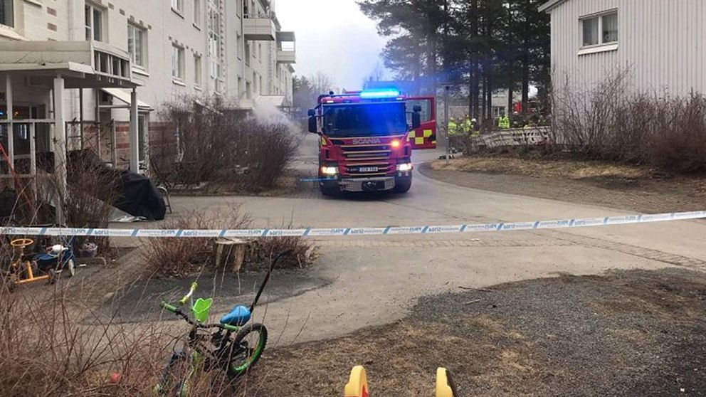 Brandbil med blinkande blåljus är parkerad på en asfalterad väg mellan vita byggnader, med rök synlig i bakgrunden. Området är avspärrat med polisband, och en barncykel ligger på marken nära avspärrningen.
