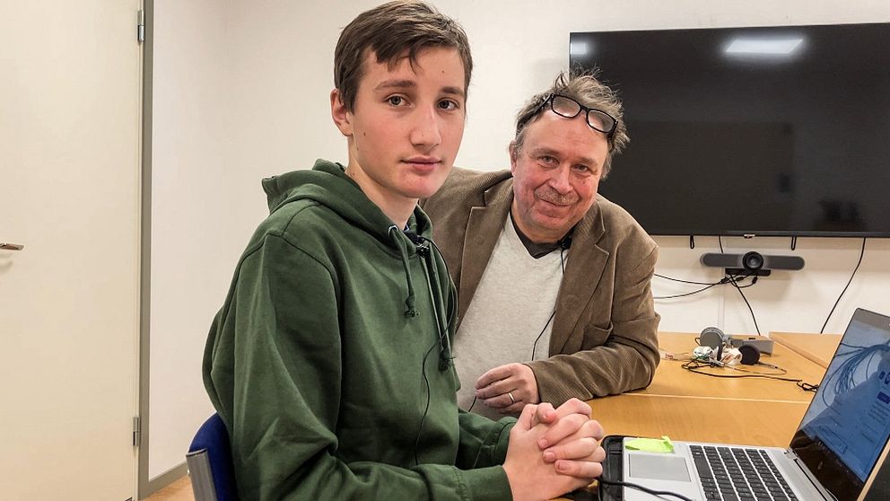 Gymnasieelven Elliot Sandström sitter i en lektionssal med knäppta händer i en grön tröja, bakom honom sitter it-pedagogen Conny Lindgren med glasögon och kavaj.