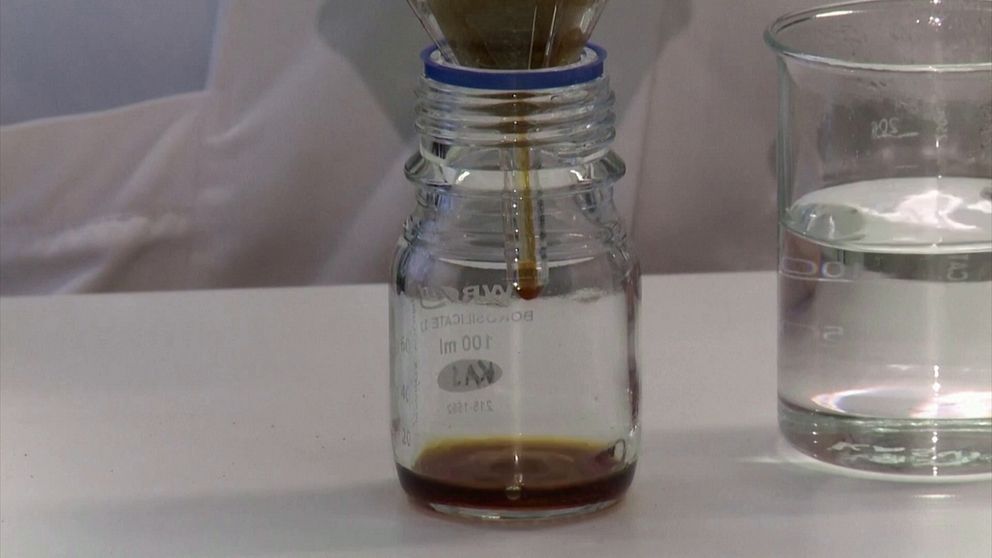 En flaska med kaffe genomgår en filtreringsprocess med hjälp av ett filter placerat ovanpå. Bredvid finns en bägare med klar vätska, allt på ett labb-bord.