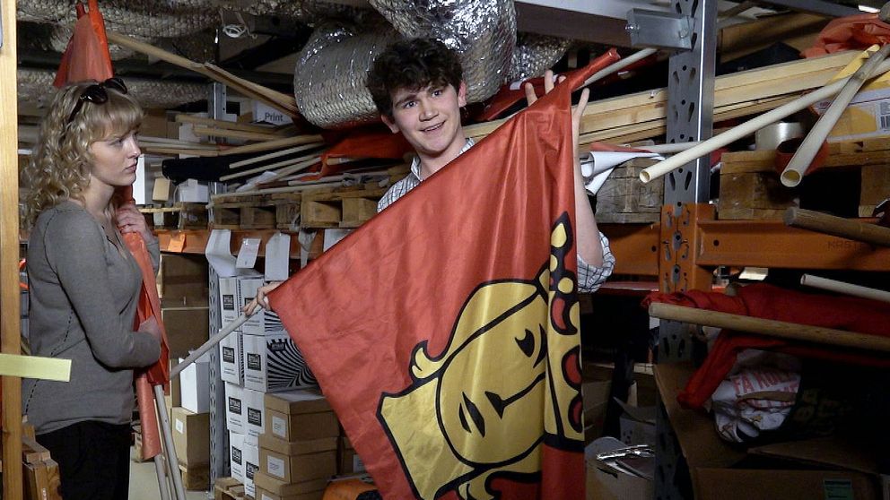 Två personer står i ett lager och håller i en röd flagga med ett gult ansikte.