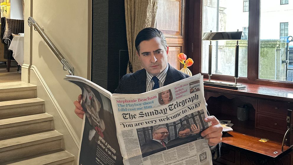 Person i kostym läser The Sunday Telegraph, står vid ett fönster nära en trappa.
