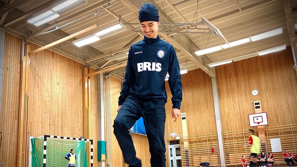 En person står i en sporthall och tränar fotboll, med ena foten som stöttar en fotboll på golvet. I bakgrunden syns en grön målvaktsbur och flera spelare som övar. Väggarna är av trä och rummet har god belysning.