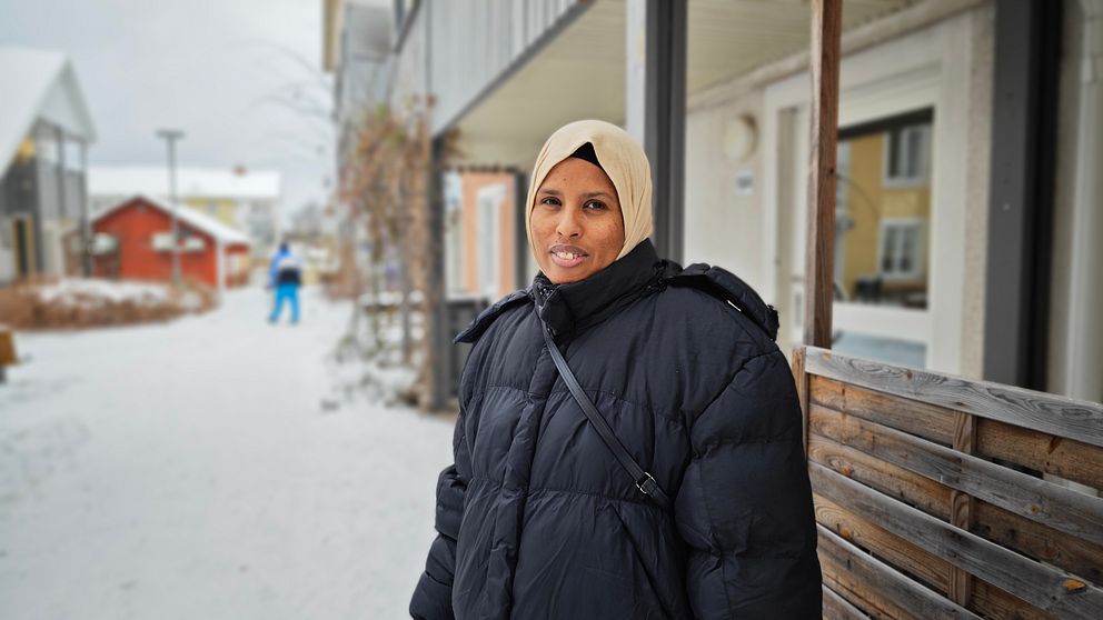 En kvinna med slöja och svart täckjacka står på en snöig innergård i ett hyreshusområde
