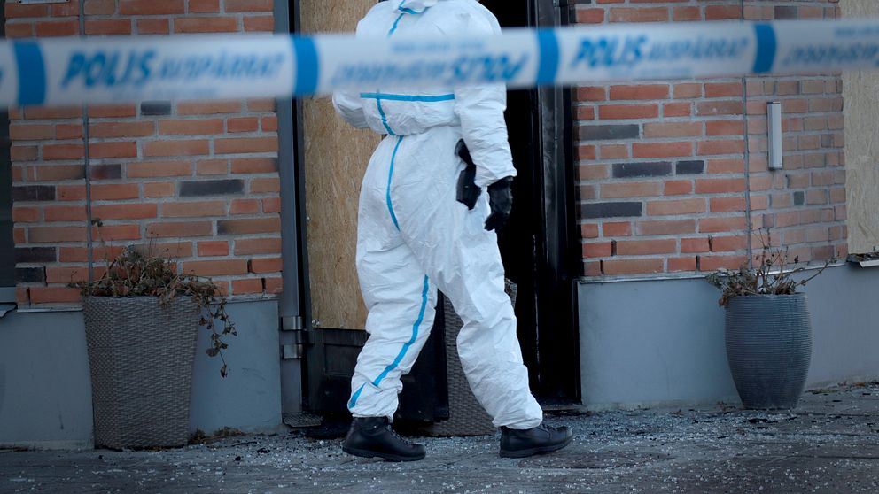En polis i skyddskläder går in genom en dörr med en uppfylld vägg av plywood. Området är avspärrat med polisband och det finns krossat glas på marken.