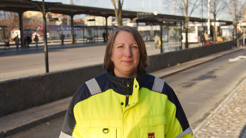 En person klädd i en neonfärgad arbetsjacka med reflexdetaljer står vid en busshållplats. I bakgrunden syns en stadsmiljö med bussar och andra människor som väntar.