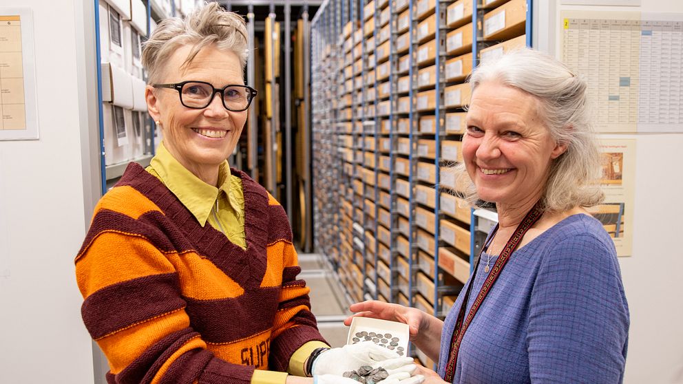 Arkeologerna Kristina Jansson och Anna Ödéen håller tillsammans i silvermynt. Bakom kvinnorna är en vägg med flera lådor.