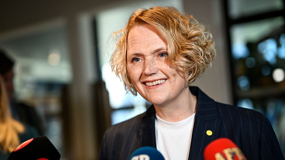 Person intervjuas av flera journalister med mikrofoner från olika nyhetsmedier.