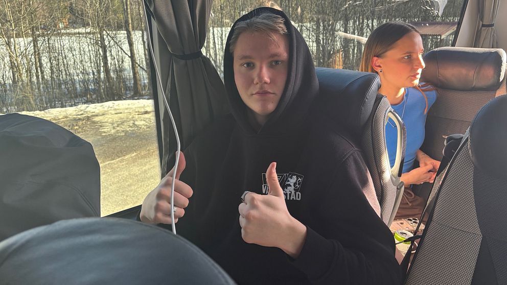Två personer på buss, en ger tummen upp. Skog ses genom fönstret.
