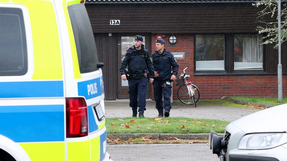Två poliser i uniform går nära en byggnad. I bakgrunden syns en cykel, och i förgrunden står en polisbil.