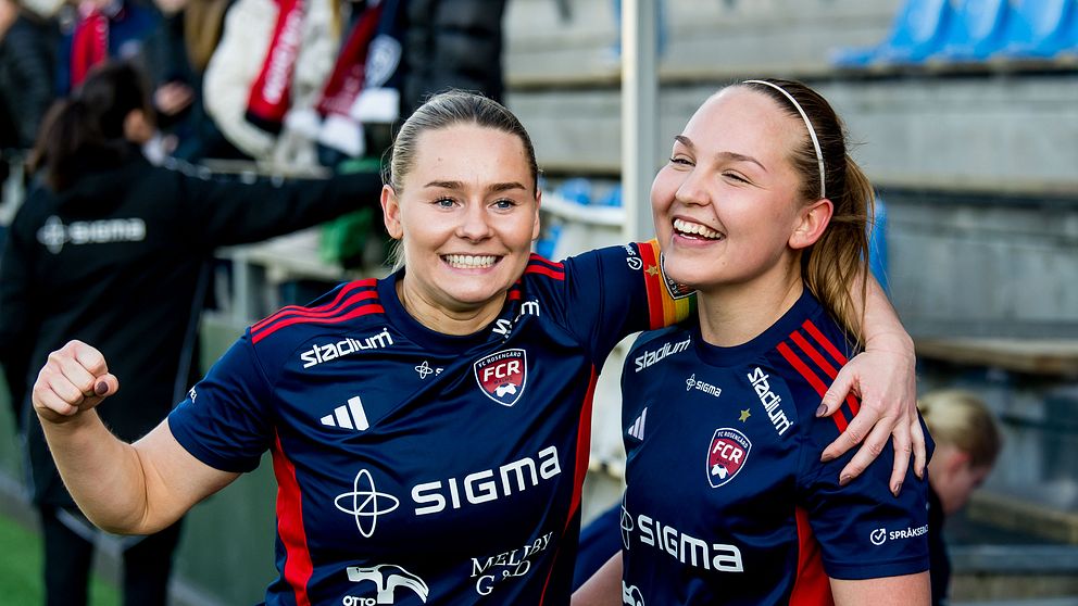 Två fotbollsspelare i blå matchställ firar tillsammans på en fotbollsarena.