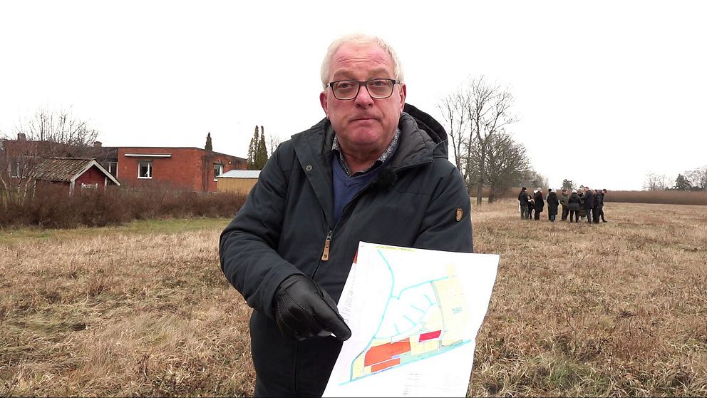 En person står utomhus på en gräsbevuxen plats, klädd i mörk jacka och svarta handskar, och håller upp en papperskarta. I bakgrunden syns en röd byggnad och en grupp människor på fältet.