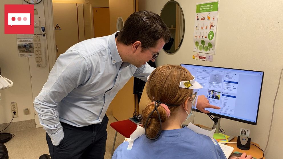 Två tandläkare står vid en dator i ett vårdrum. Läkaren pekar på skärmen medan sjuksköterskan följer hans anvisningar. Datorn visar en medicinsk presentation eller journal, vilket indikerar diskussionen om patientvård. I bakgrunden syns sjukhusmiljö och medicinska affischer.