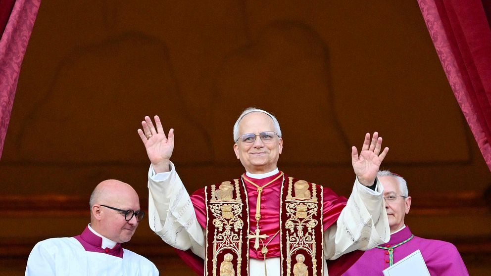 Påve står på en balkong med händerna upplyfta, flankerad av präster i liturgiska kläder.