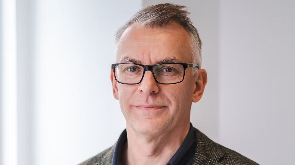 En man står framför ett ljust rum, iklädd en mönstrad kostymjacka över en mörk skjorta. Bakgrunden är ljus och neutral.