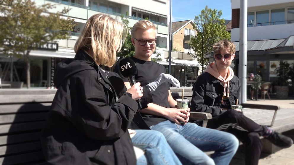 Reporter intervjuar två personer på bänk utomhus i stadsmiljö.