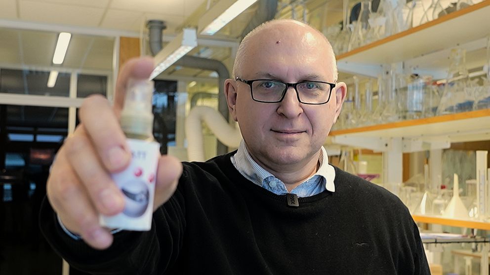 Person i labbrock håller en liten flaska i ett laboratorium fullt med utrustning och glasbehållare i hyllor.