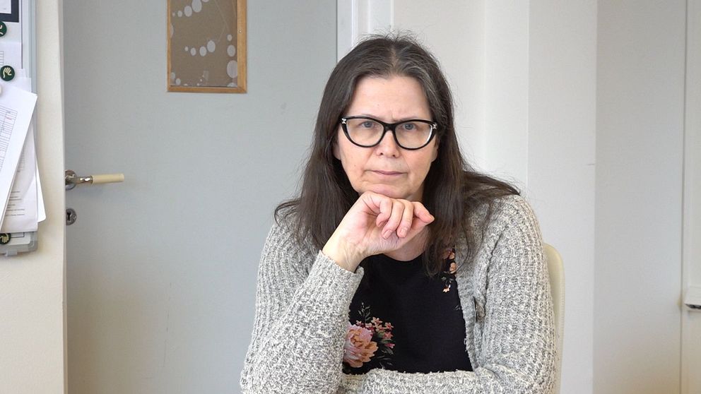 En person sitter i ett kontorsutrymme med papper och en anslagstavla i bakgrunden. Personen bär en stickad tröja.