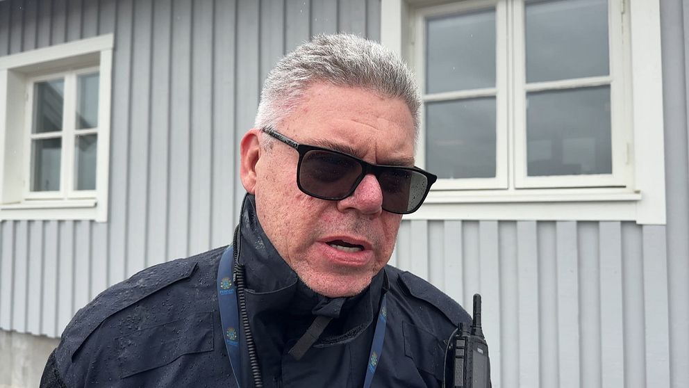 Person med radiokommunikationsutrustning framför grått hus med vita fönster.
