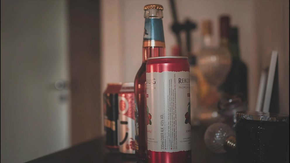 En uppsättning ciderburkar och flaskor på ett bord som inkluderar en röd burk med vit text i förgrunden, en flaska öl med en blårandig kapsyl bakom den, och en annan burk bakom den med ett mönster. I bakgrunden syns suddiga flaskor av olika slag.