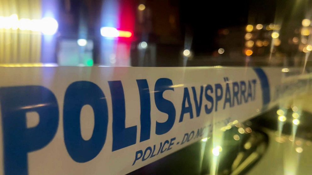 En närbild av ett avspärrat område med en polisavspärrningsbanderoll som visar texten ”POLIS AVSPÄRRAT” och ”POLICE DO NOT CROSS”. I bakgrunden syns suddiga ljus från polisfordon.