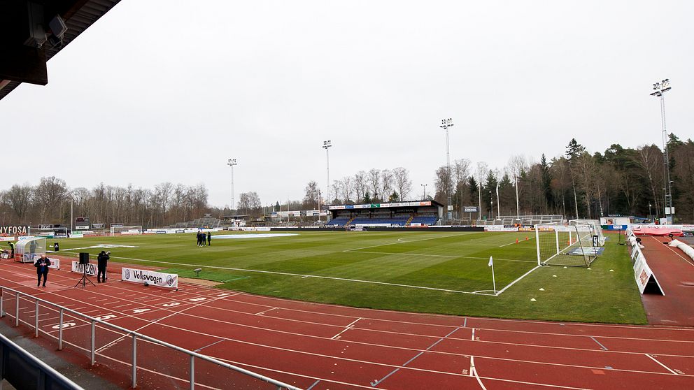 En tom fotbollsarena med gräsplan och löparbanor under en grå himmel.