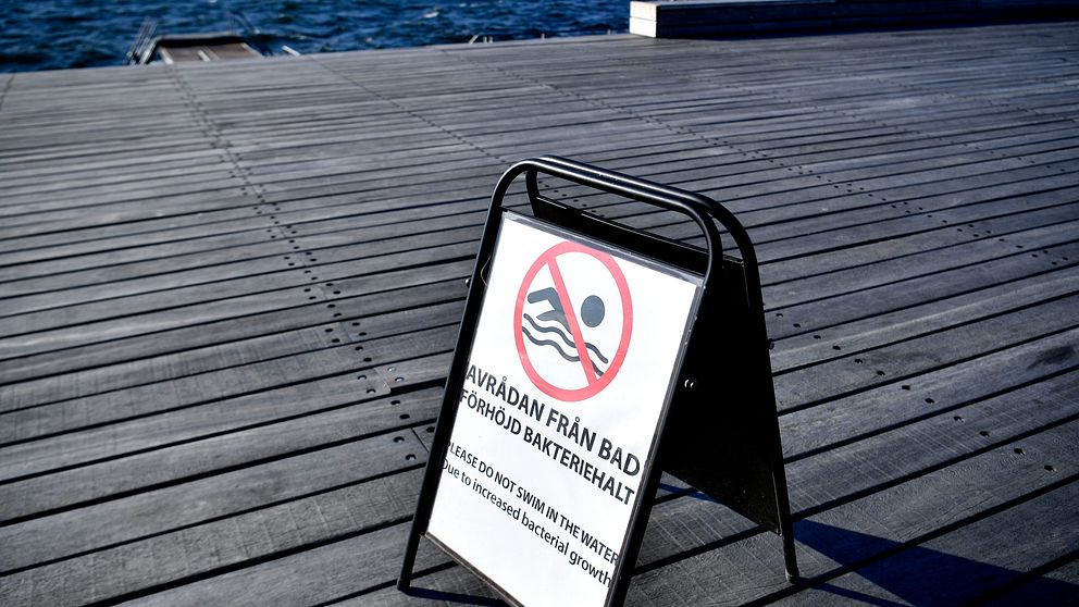 Skylt med texten ”Avrådan från bad – förhöjd bakteriehalt” uppställd på trädäck, vid vatten.