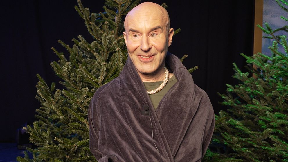 En person i en mörk badrock står framför ett par granar på en scen med svag belysning.