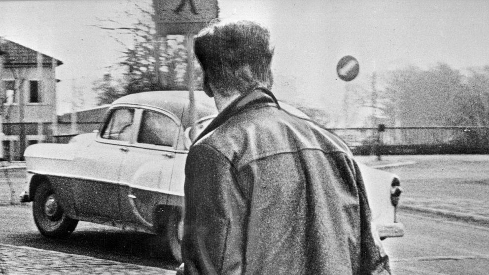 Ung man med pistol går på trottoar, vintagebil i bakgrunden.