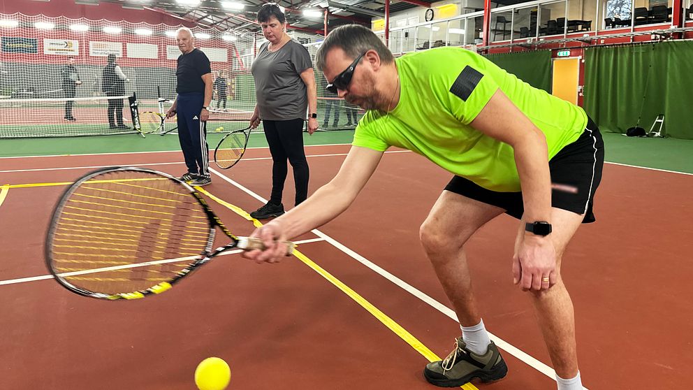 En tennisövning på inomhustennisbana där en synskadad spelare i grönt tränar sin teknik genom att slå en tennisboll med racketen, medan två andra spelare observerar i bakgrunden.