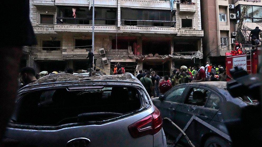 Räddningspersonal arbetar vid en skadad byggnad efter en explosion. I förgrunden syns två skadade bilar, med en av dem med trasig bakruta. Bakom dem är en skadad byggnad med flera öppningar och människor som evakuerar området. Räddningsfordon är synliga i bakgrunden.