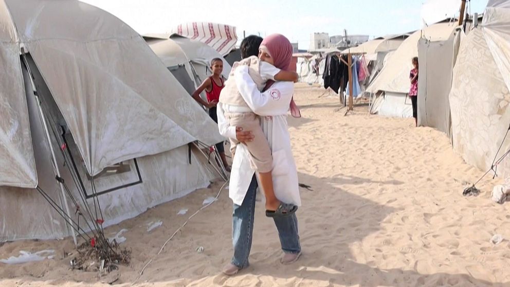 En sjukvårdare i Gaza bär ett barn genom ett flyktingläger med tält på båda sidor. Sandig mark syns mellan tälten, där flera personer är synliga i bakgrunden. Atmosfären tyder på en situation med humanitärt stöd.