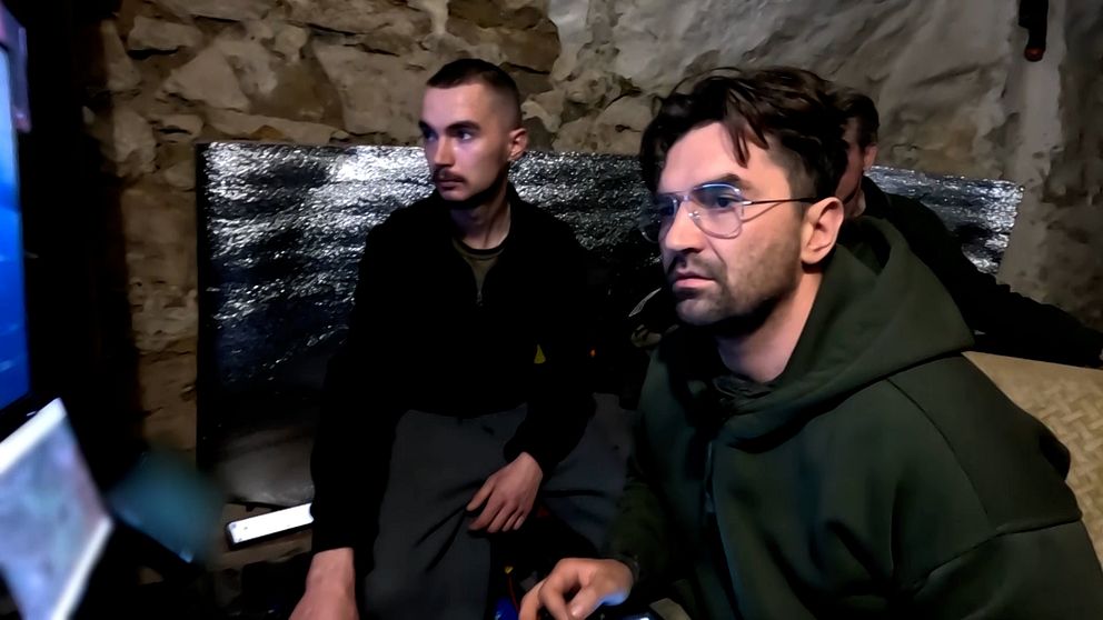 Två personer sitter framför drönar-skärmar i ett rum med stenmur och isolering.