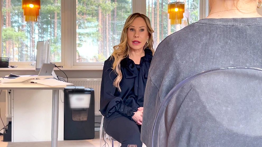 En blond kvinna sitter framåtlutad i en modern inomhusmiljö, med ett skrivbord i bakgrunden. Reportern sitter i en stol mittemot med ryggen mot kameran.