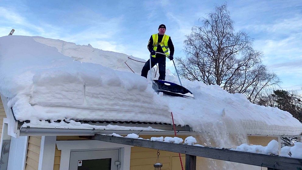 Man med varselväst står med en snösläde på ett villatak med snö.