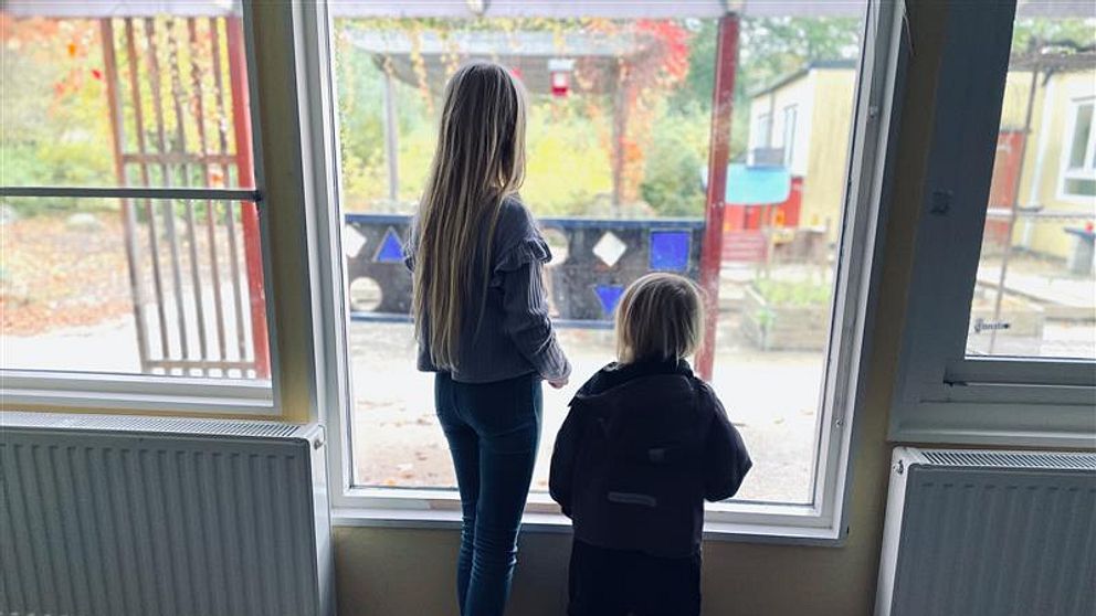 Två barn står vid ett fönster och tittar ut. Den äldre flickan har långt hår och står barfota, medan den yngre flickan är klädd i en overall. Utsikten visar en lekplats med färgglada leksaker i bakgrunden och höstlöven syns utanför.