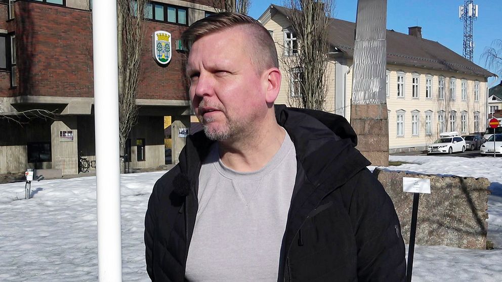 Haparandas kommunalråd Janne Lind står utomhus utanför Haparanda stadshus. Det är soligt och hans svarta jacka är uppknäppt .