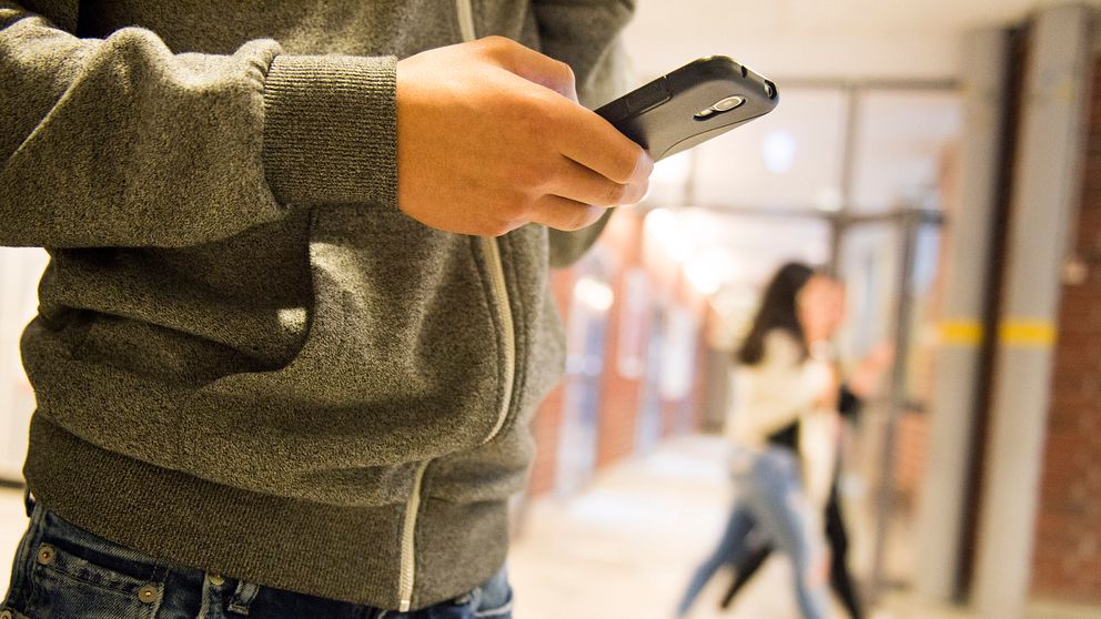 En person i grå hoodie håller en mobiltelefon i handen, medan två personer går förbi i bakgrunden i en skolmiljö.