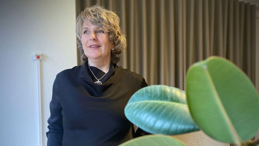 Pia Idhede Liljegren står i en inomhusmiljö iklädd en svart tröja, riktad mot kameran. I förgrunden syns gröna blad från en växt vilket ger en känsla av livfullhet i rummet. Bakgrunden består av ljusa väggar och en gardin.