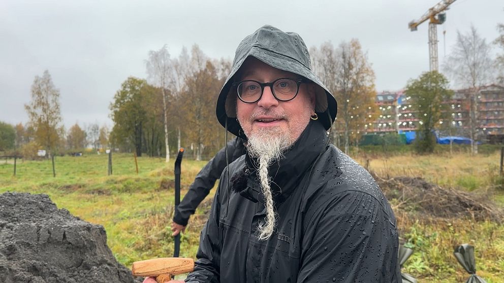 En person i regnkläder står i en fältmiljö, omgiven av träd och byggnader i bakgrunden. Det regnar och marken är blöt, medan personen håller i en träspade och fyller sandsäckar med sand.