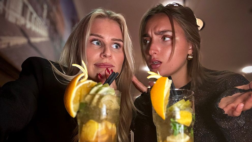 Två personer poserar med varsin drink på ett bord med röd-vitrutig duk. Drinkarna är dekorerade med citrusskivor och myntablad, vilket ger en livlig och uppfriskande känsla. I bakgrunden syns en suddig interiör av en restaurang.