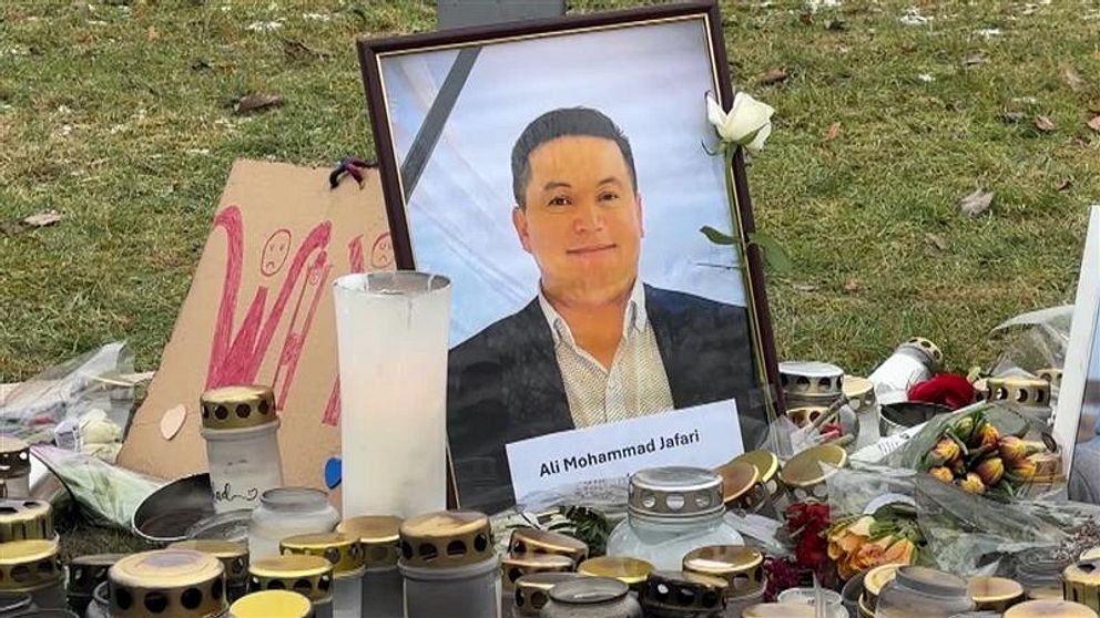 Minnesplats utomhus med ett inramat fotografi av en man, omgiven av tända ljus, blommor och en skylt med namnet ”Ali Mohammad Jafari”.