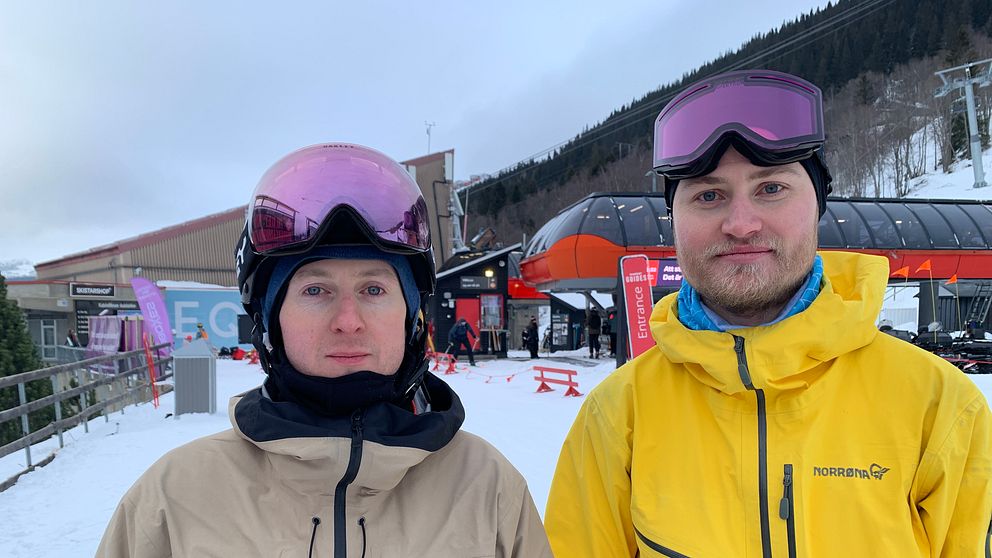 Två personer klädda i skidkläder står vid en skidort. Personen till vänster bär en beige jacka och ett rosa visir, medan personen till höger har en gul jacka och ett mörkt visir. I bakgrunden syns en skidlift och byggnader omgivna av snö.
