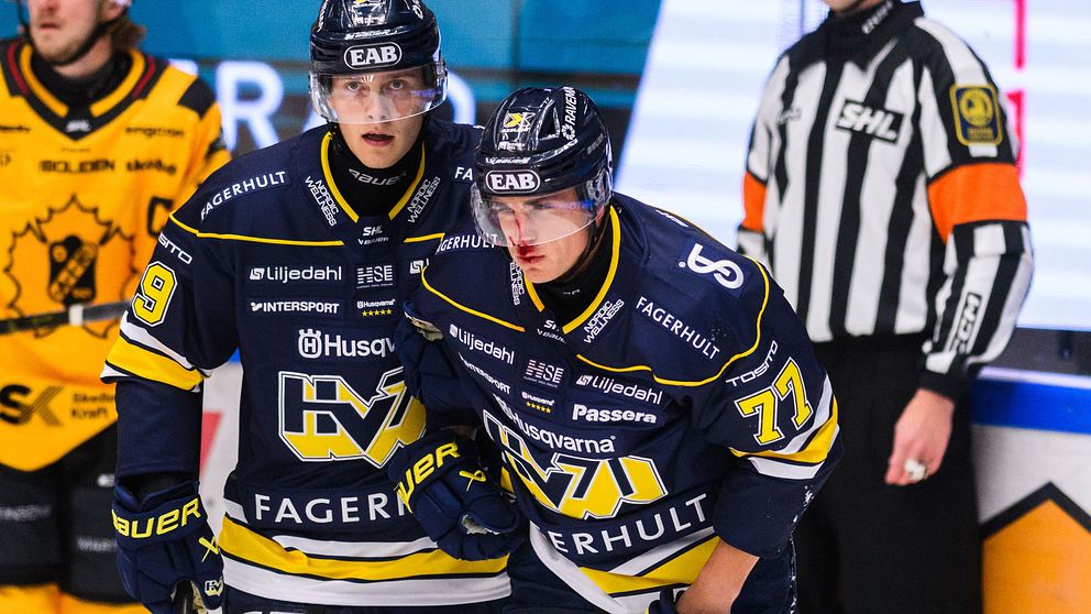 Två ishockeyspelare i matchtröjor från HV71 står tillsammans på isen. Den ena spelaren, Herman Hansson, ser ut att ha ont. I bakgrunden syns en domare i svart och vitt, och det är en energisk atmosfär i rinkens miljö.