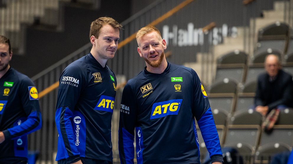 Två handbollsspelare i mörkblå träningskläder pratar på läktare inför match.