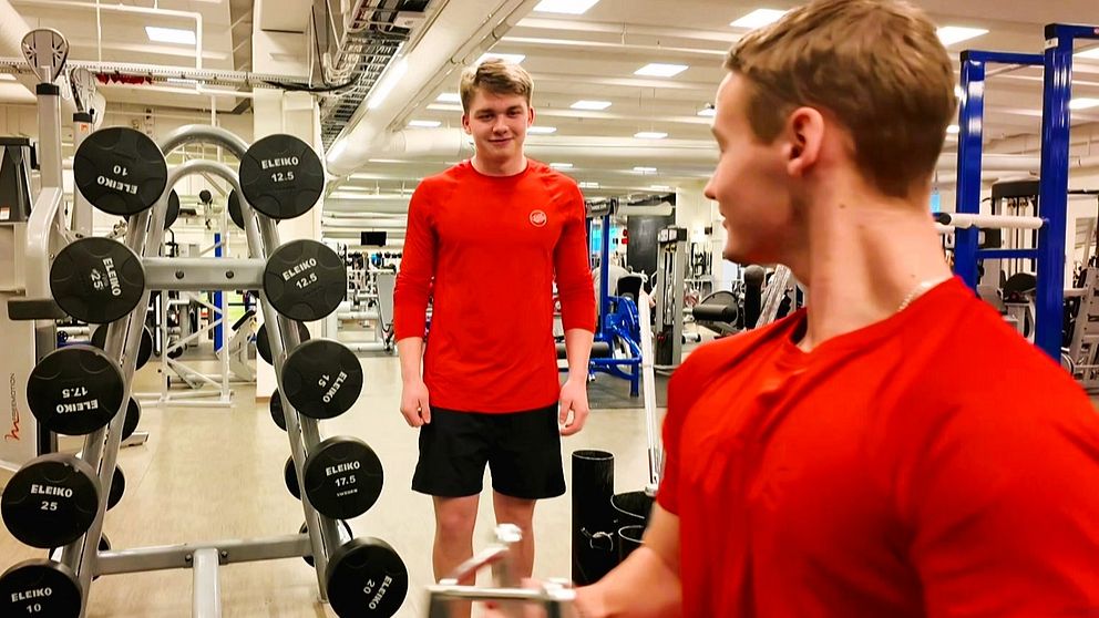 Två killar på ett gym, den ena drar i ett handtag för ryggträning medan hans kompis tittar på.