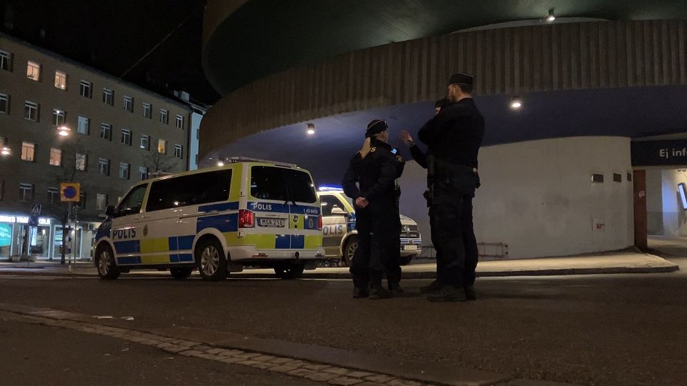 Två poliser står på en gata om natten i närheten av en polisbil, medan en byggnad med rundad struktur syns i bakgrunden.
