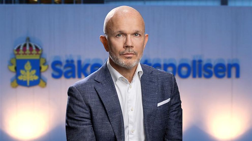 En person i kostym står framför en bakgrund med texten ”Säkerhetspolisen” och säpos logotyp. Belysningen är mjuk och fokuserar på personen.